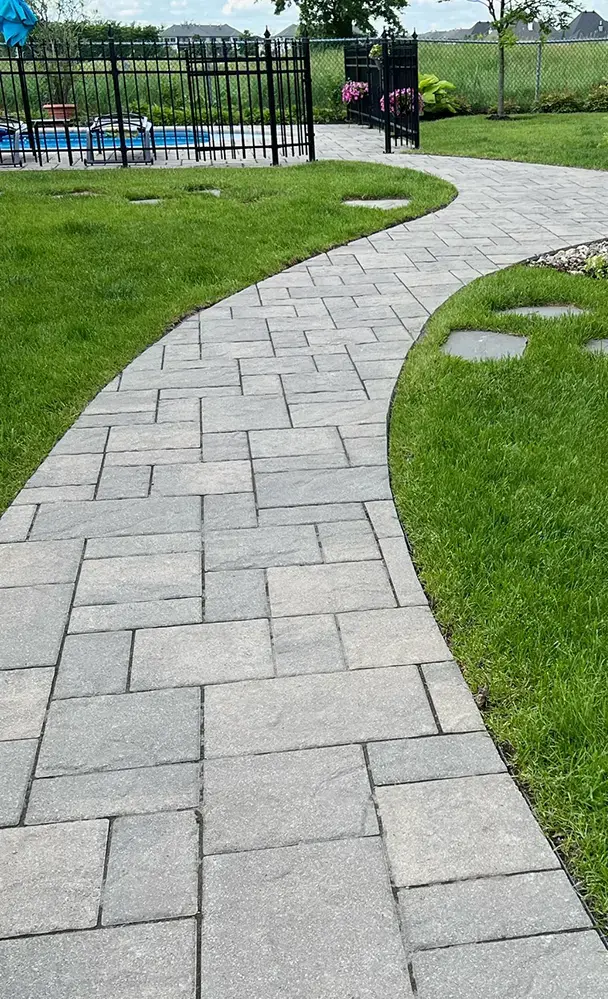 Chemin en pavé uni sinueux menant à une piscine, entouré de pelouse verte et d’une clôture noire, dans un aménagement paysager résidentiel.