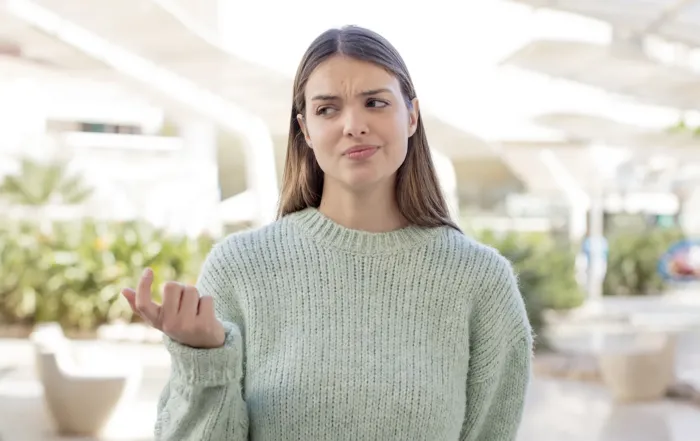 Femme pensive en chandail vert pâle, regard interrogateur, à l’extérieur dans un environnement urbain avec de la verdure.